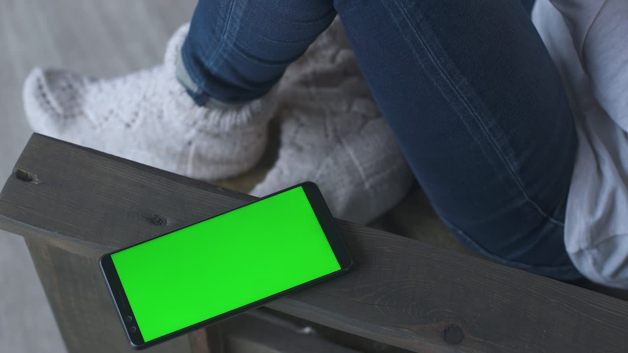 짧은 머리카락과 청바지를 입은 소녀는 집에서 의자에 앉아서 겨울에 책을 읽습니다. 초록색 화면의 휴대 전화 근처에. 휴대 전화를 가진 여자. 여성 휴대전화. 크로마 키 휴대폰 녹색 화면.
