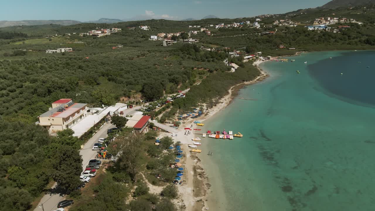 Resort buildings and beach umbrellas line the shallow turquoise coast of Lake Kournas in Crete with pedal boats and forested countryside visible in the background