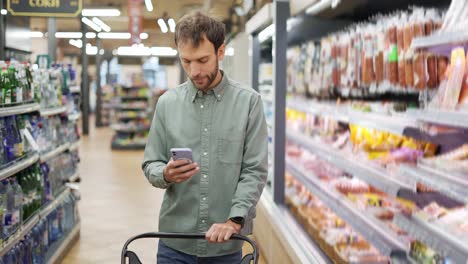 happy man in shirt walking with a cart through the supermarket choosing groceries. checking the shopping list on the cell phone