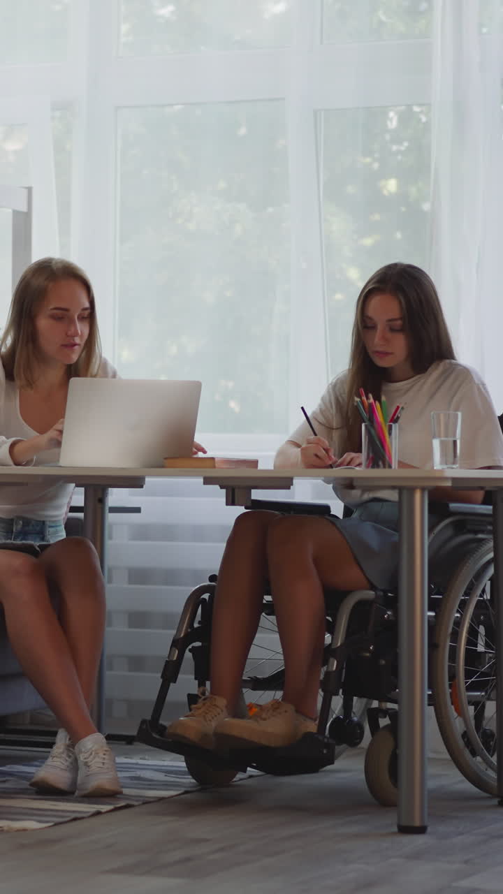 las mejores amigas hacen la tarea juntas sentadas en la mesa. la mujer rubia usa la computadora portátil mientras que la dama que usa la silla de ruedas hace notas en el bloc de notas