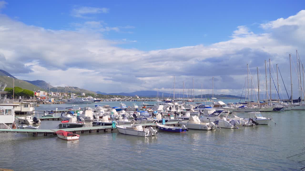 Vista panoramica di motoscafi e barche a vela bianchi ormeggiati a pontili galleggianti al porto turistico di Gaeta con catena montuosa sullo sfondo in una giornata di sole, Italia, statica.