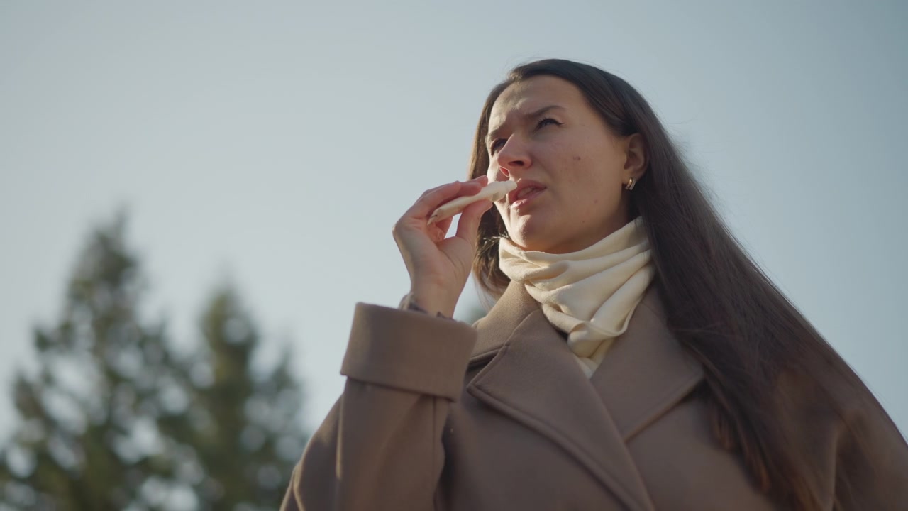 Primo piano di una donna che tiene e usa un balsamo labbra all'aperto con alberi sullo sfondo. La donna indossa un cappotto marrone e una sciarpa color crema, mentre dimostra l'applicazione del prodotto su uno sfondo di cielo sereno.