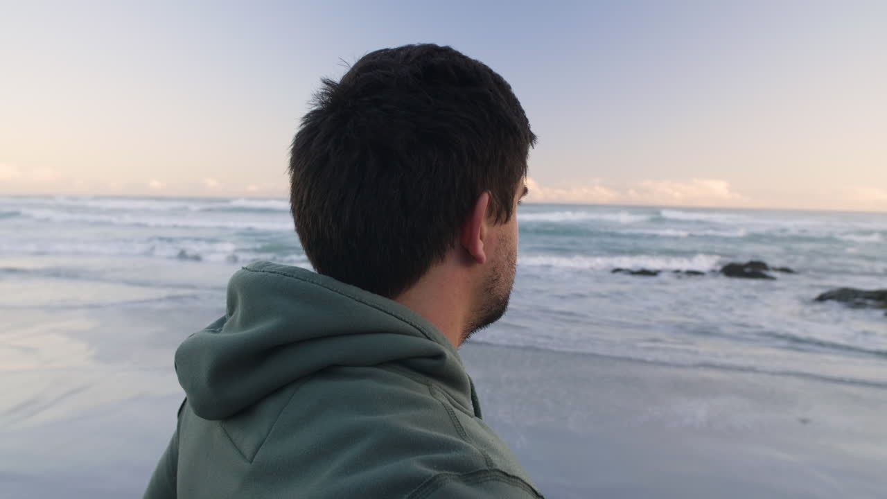 Camera orbits around young male beach jogger in green hoodie, ending in left of frame as he stares out to the ocean in early morning sunlight