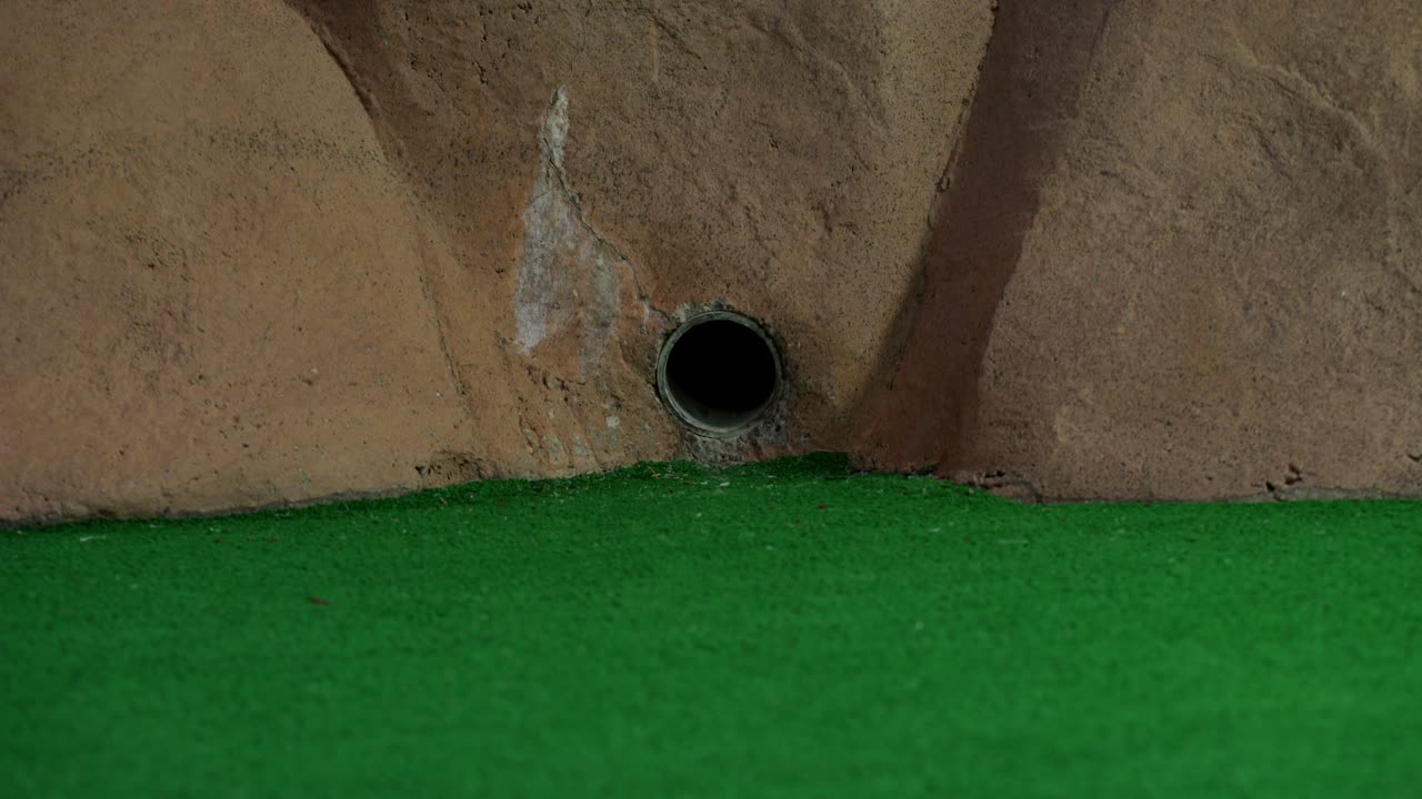 una pelota de minigolf naranja sale de una tubería en una pared y rueda y no alcanza el hoyo
