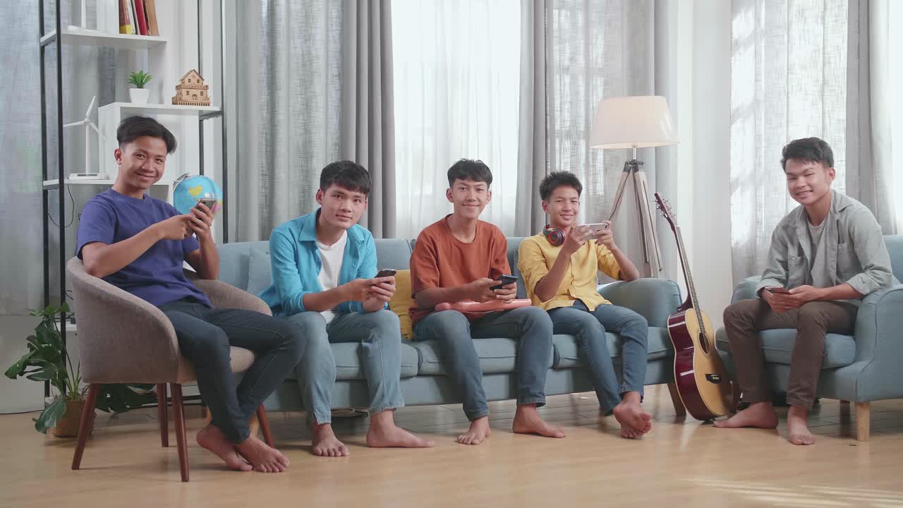 젊은 아시아 친구 그룹이 악기를 들고 집에서 소파에서 스마트폰을 재생하는 동안 미소 짓고 머리를 카메라로 흔들고 있습니다.