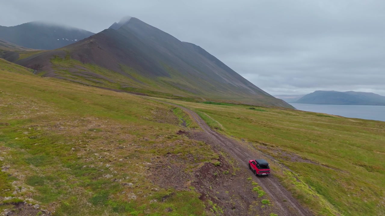 Una ripresa aerea di un'auto 4x4 rossa che viaggia lungo la remota strada Svalvogaviti in Islanda, con imponenti cime montuose e viste costiere.