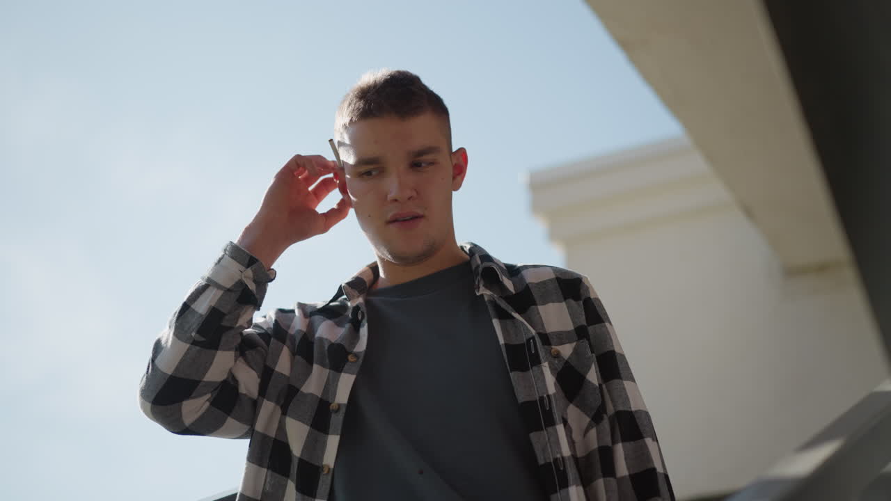 young man in black white check flannel shirt descending outdoor metal staircase grips cigarette from behind ear raises hand flicks lighter ignites tip with bright spark then prepares inhale smoke