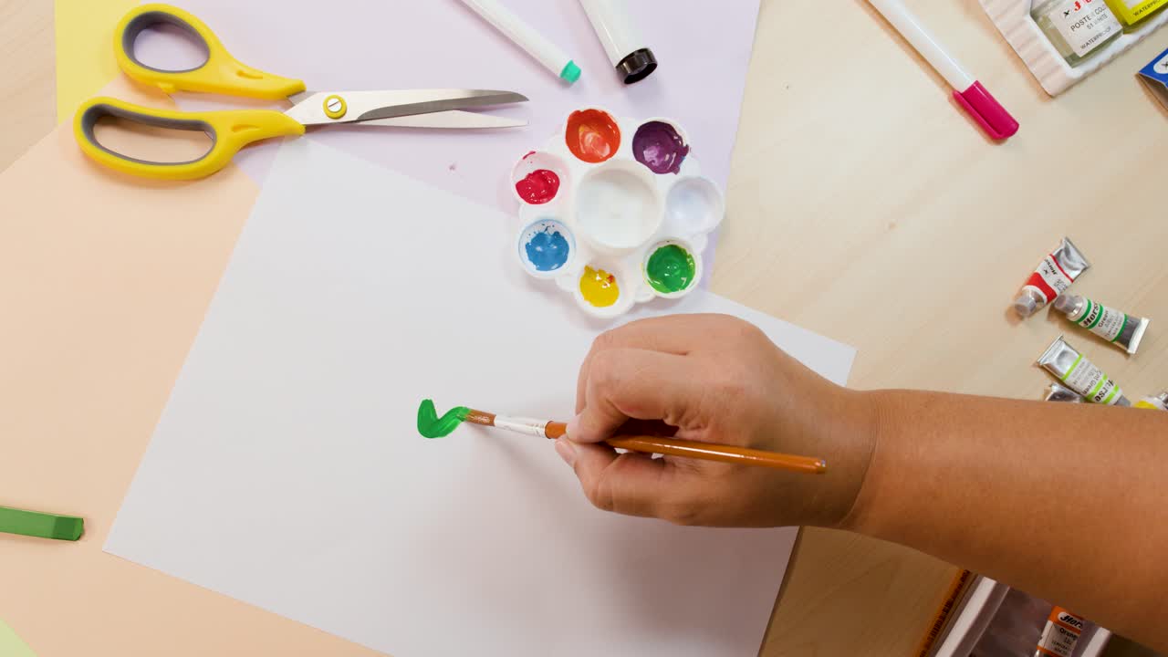 Overhead view of hand painting green wavy line on white paper with acrylics, bright lighting