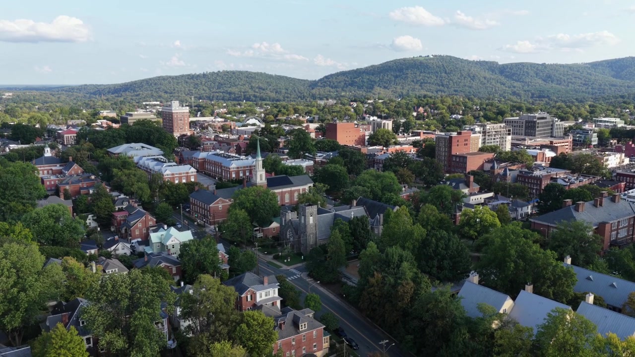 Luce calda serale su Charlottesville, Virginia, con tetti storici, isolati del centro, fitta vegetazione di fine estate e la lunga cresta boscosa delle Blue Ridge. Ripresa aerea panoramica con drone