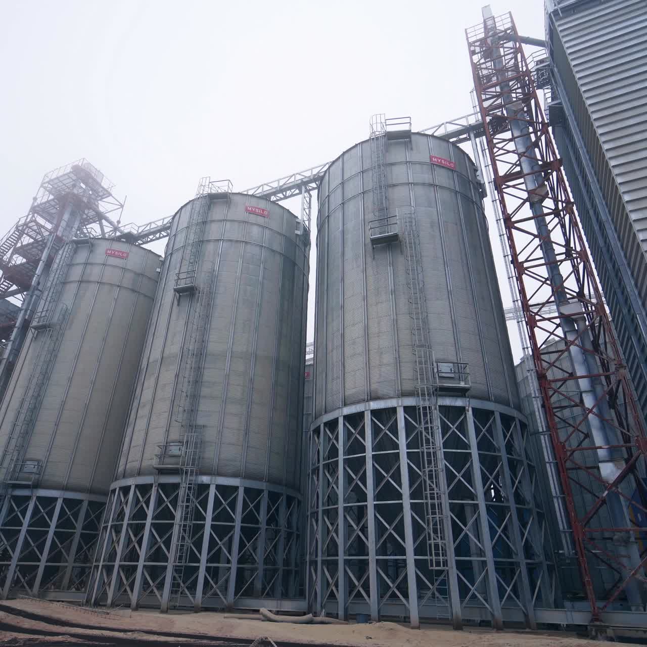 Three big silver cisterns standing in row on steel supports. Lorry standing under the grain storage elevator. High silo constructions against the clear sky