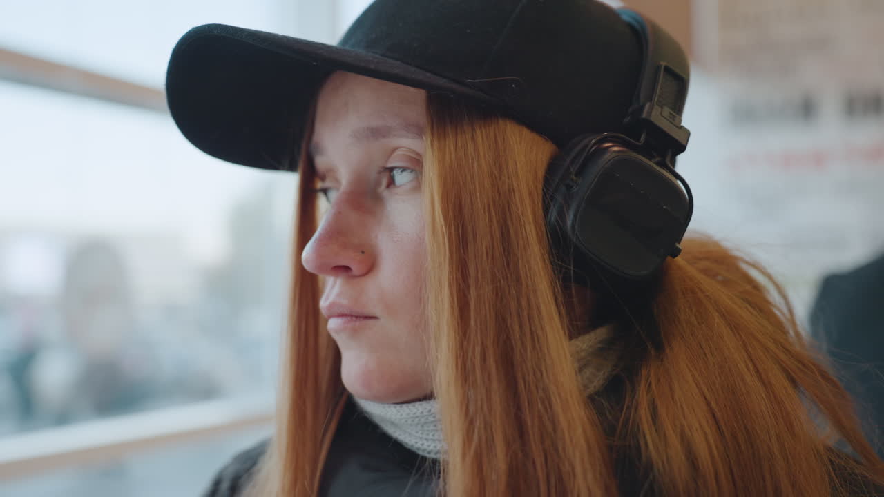 駅で乗り物の窓から外を見ているヘッドホンをつけた若い女性。黒いキャップの下に見える赤毛、かすかなそばかす、重ね着のコートとパーカー、物思いにふける表情。クローズアップの連続が変化を示している。