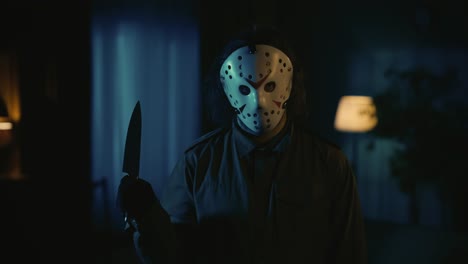man maniac in white hockey mask standing in the room looking directly at the camera and holding