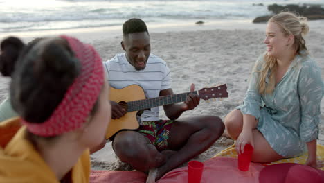 Un-Joven-Afroamericano-Toca-La-Guitarra-En-La-Playa-En-Una-Fiesta,-Rodeado-De-Amigos.