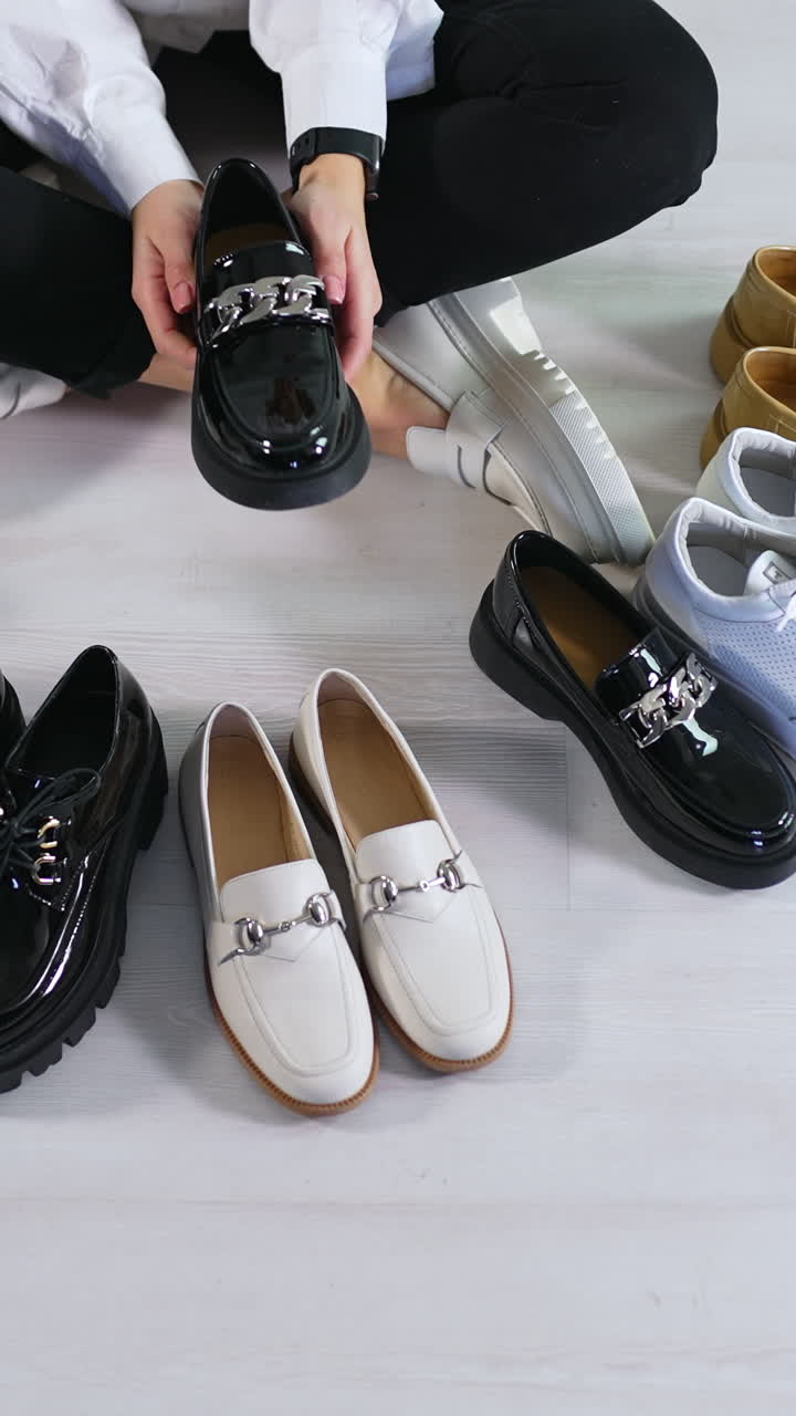 New pairs of modern footwear on the floor in front of sitting lady. Woman runs her hands over the diverse shoes choosing one black lacquered loafer. View from above. Vertical video