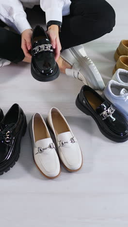 new pairs of modern footwear on the floor in front of sitting lady. woman runs her hands over the diverse shoes choosing one black lacquered loafer. view from above. vertical video