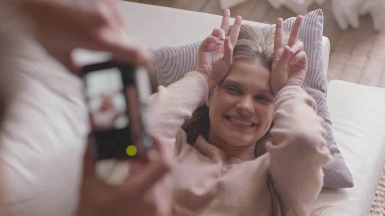 행복한 젊은 커플이 카메라를 사용하여 함께 사진을 찍고 소파에서 집에서 재미있게 로맨틱한 관계를 즐기며 서로 사진을 찍고 얼굴을 짓고 있습니다.