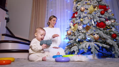 happy caucasian woman decorating christmas tree. lovely baby sitting beside plays with toys. low angle view.