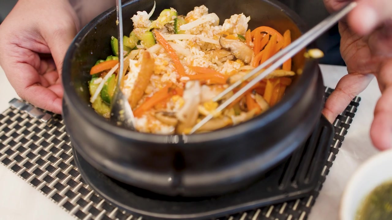 Close-up de mãos misturando bibimbap em uma tigela de pedra ao lado de sopa de algas e kimchi.