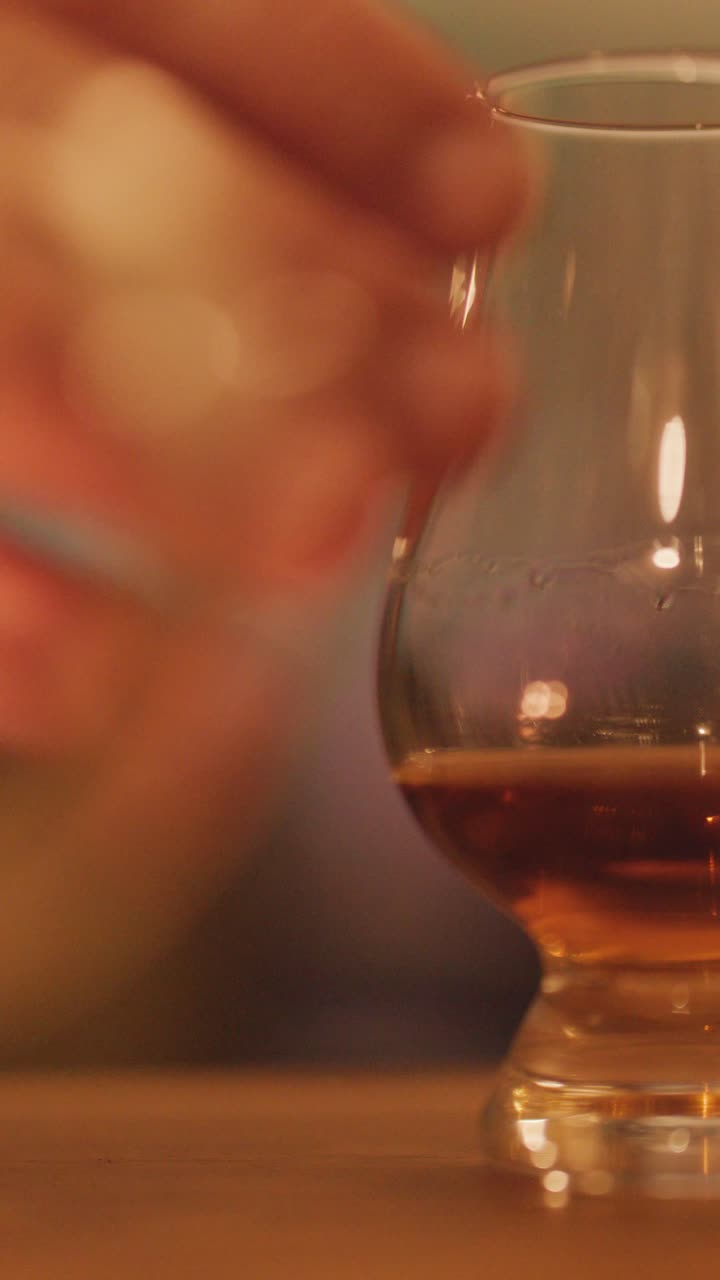 Una mano si allunga per prendere e solleva un bicchiere di whisky da un tavolo di legno in un bar illuminato soffusamente con sfondo sfocato e un'atmosfera calda a lume di candela