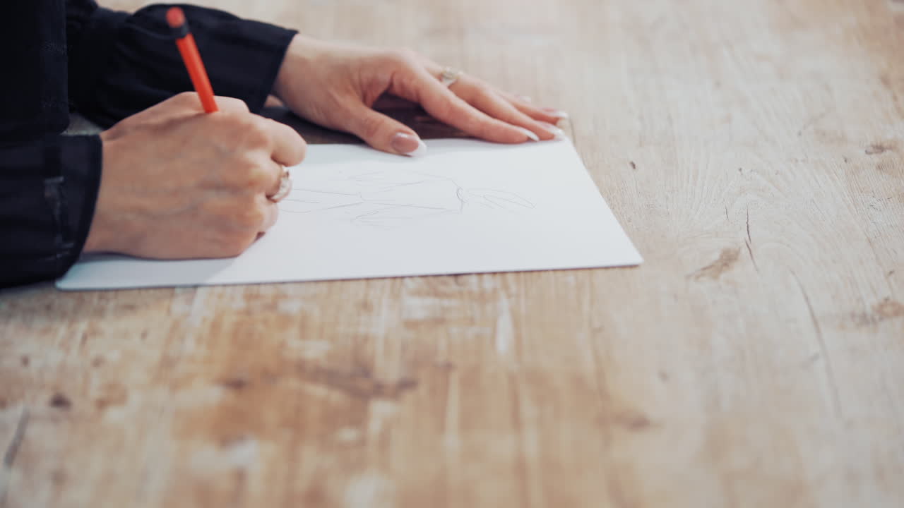 Dressmaker is drawing a creative design of a modern clothing indoors. Female's hands of a young tailor on the wooden table with a pencil. Feminine hobby.