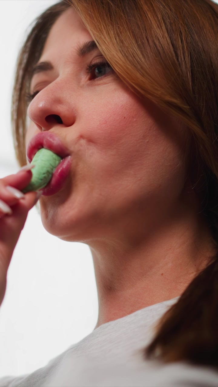 Close up of woman eating green ice cream with her lips visible. subject holds cone near their mouth wearing white top against plain background. Soft lighting highlights facial features