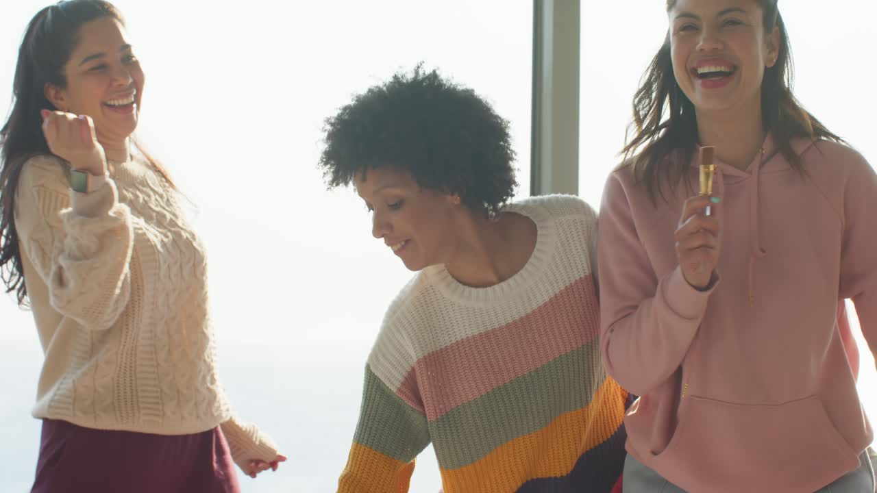 Diverse female friends shaking bottle grabbing snack bar and dancing for morning boost at window