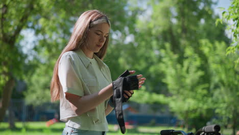 young woman standing outdoors, thoughtfully removing biker glove from right hand, with lush greenery and trees in blurred background, background includes rail and park elements