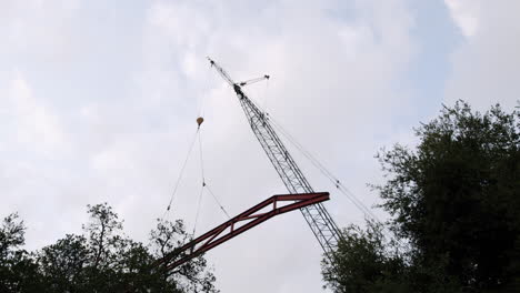 a large crane lifting a red metal truss structure against a cloudy sky, with trees in the foreground
