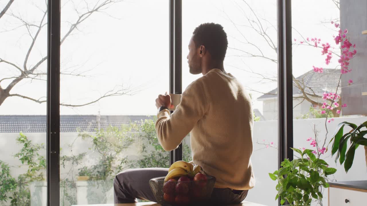 Happy african american man sitting at countertop in kitchen, drinking coffee
