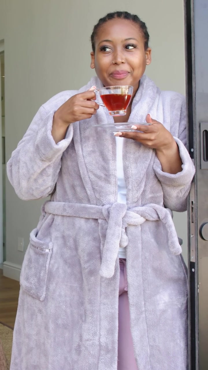 Vertical video: Lifting glass teacup, woman in robe sipping tea in doorway holding saucer for calm