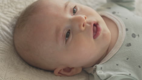 cute happy baby laying on his back in white bed sheets, looking with curiosity, portrait in bright bedroom