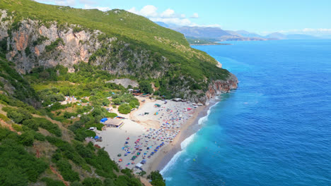 aerial drone zoom in shot of tourists sunbathing along the gjipe beach in albania with waves crashing along the beachside