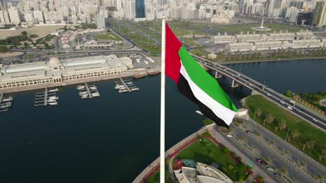 Einzigartige-Luftaufnahme:-Nationalflagge-Der-Vereinigten-Arabischen-Emirate-Weht-Im-Wind-über-Der-Flaggeninsel-Von-Sharjah,-Sharjah-Stadt-Im-Hintergrund,-Flagge-Der-VAE,-4K-UHD-60 fps