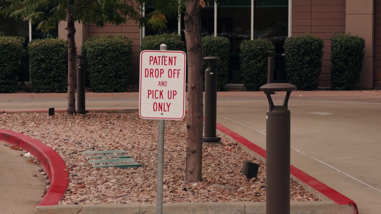 A static view centers on a patient drop-off sign beside a medical facility driveway.