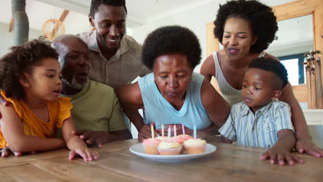 Una-Familia-Multigeneracional-Celebra-El-Cumpleaños-De-La-Abuela-Con-Pastel-Y-Velas-Alrededor-De-La-Mesa-En-Casa