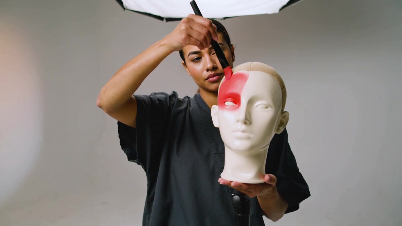 Una donna in camice scuro posiziona un manichino, applicando pigmento rosso all'occhio destro con un pennello in uno studio.
