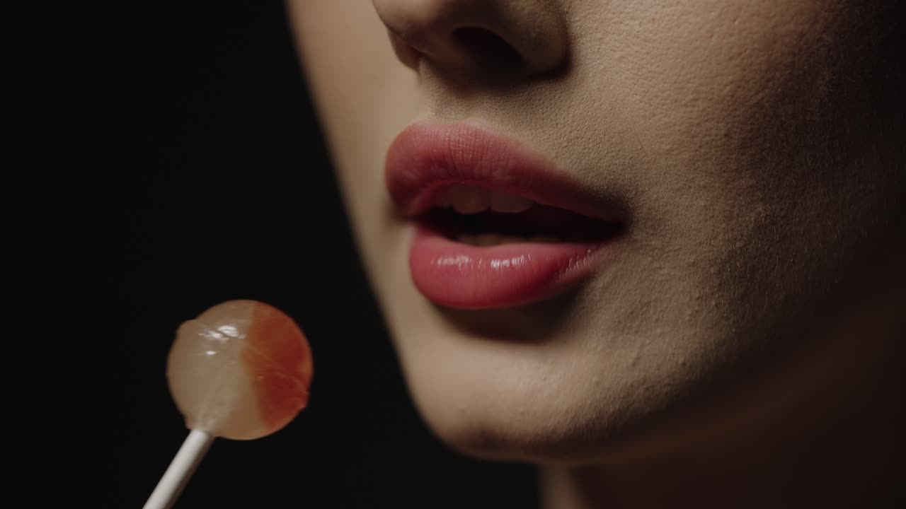 vista de cerca de una seductora rubia joven mujer caucásica con labios rojos lamiendo una deliciosa piruleta dulce y disfrutando del caramelo contra un fondo negro en cámara lenta
