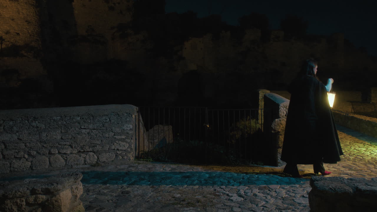 una mujer misteriosa camina de noche con una linterna cerca de un castillo.