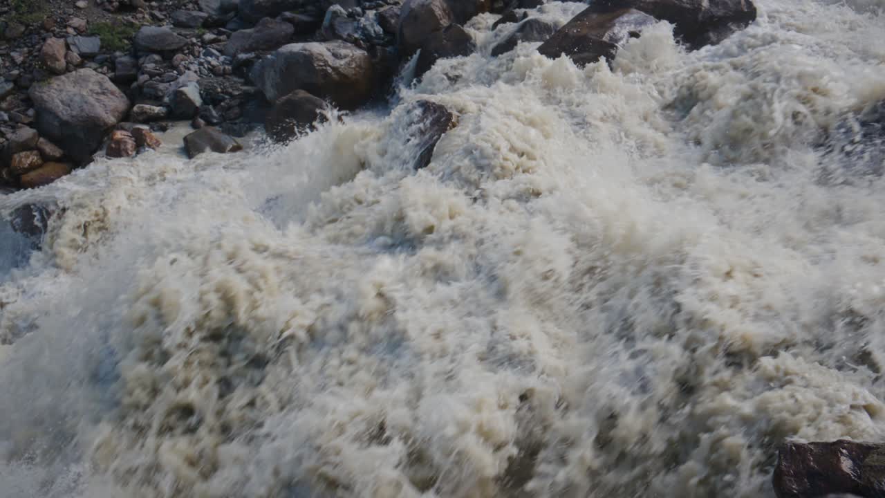 Tosenes Flusswasser schlägt über dunkle Felsen. Schaum spritzt in chaotischer Bewegung nach oben. Die Szene zeigt die rohe Kraft ungezähmter Natur. Ideal für Abenteuer-, Umwelt- oder Extremwetterthemen.