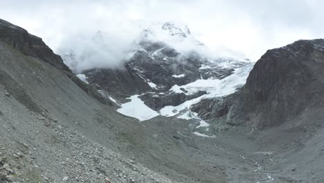 Vista-De-Drones-Sobre-Un-Desolado-Glaciar-Alpino-En-Retroceso-Rodeado-De-Montañas-Y-Rocas-Y-Picos-Cubiertos-De-Nieve