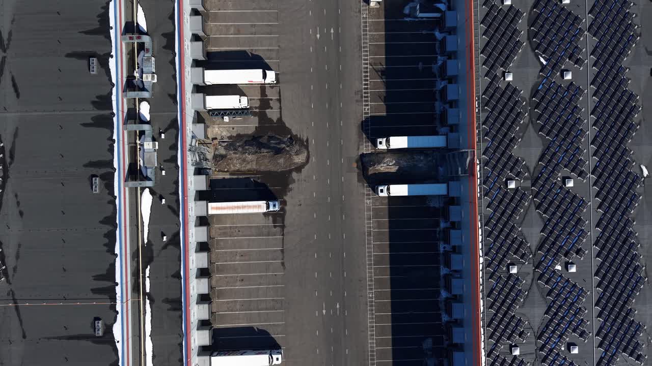 Drone shot over an industrial area with loading docks, trailers, and a warehouse. The scene captures a sunny day with clear visibility, showcasing organized structures and empty parking spaces