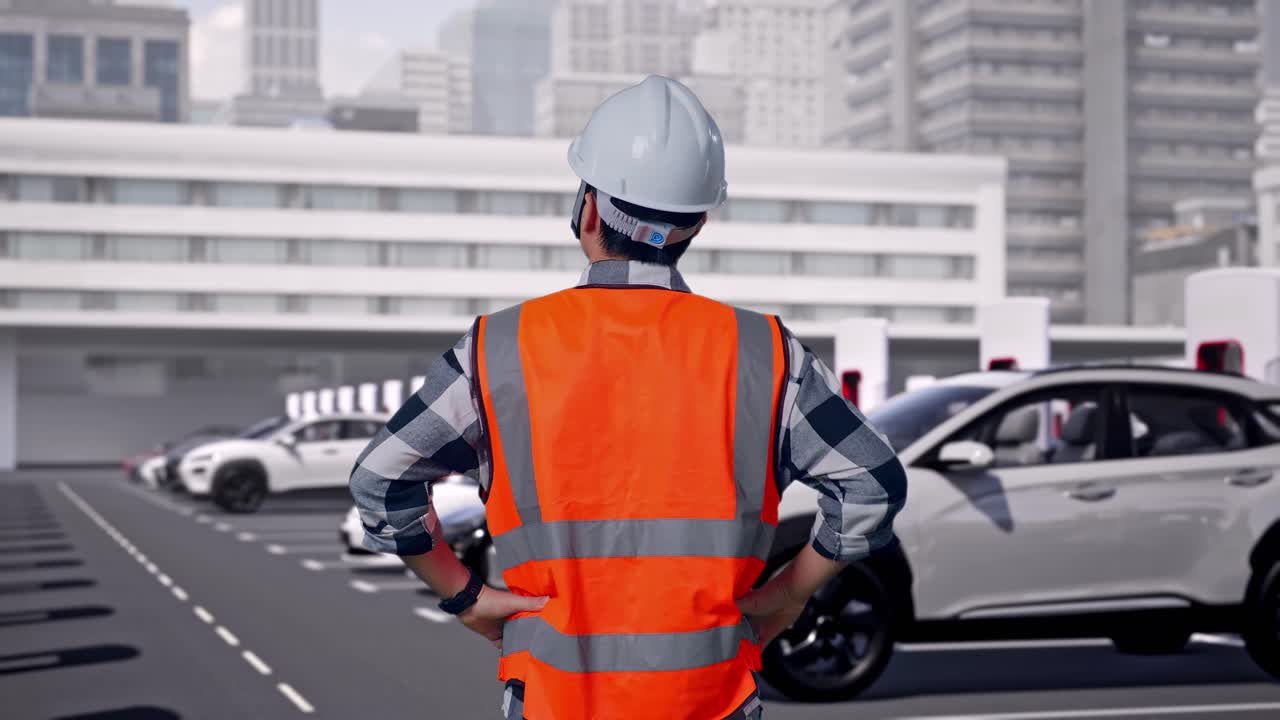 近代都市の充電ステーションで電気自動車が充電されている中、安全ヘルメットを着用した男性エンジニアが腰に手を当てて立ち、周囲を見回している後ろ姿。