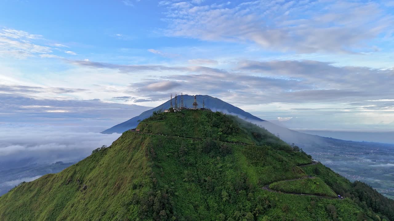 Drone terbang naik menunjukkan pemandangan puncak Gunung Telomoyo dengan latar belakang Gunung Merbabu, lanskap pedesaan Indonesia