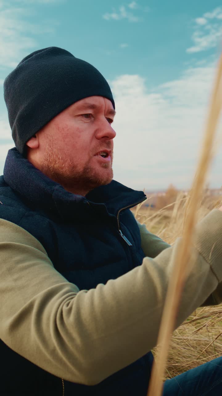 Een man met een muts en vest die een smartphone gebruikt te midden van hoog gras onder een blauwe hemel, momenten vastlegt of geniet van de natuur tijdens een ontspannende buitenervaring