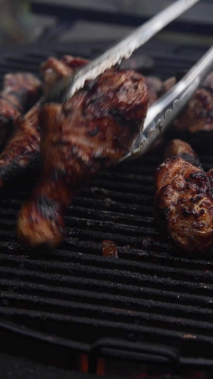 Video verticale in primo piano di cosce di pollo marinate che cuociono su un barbecue. Pinze di metallo che girano la carne succosa su una griglia affumicatrice a carbone ardente.