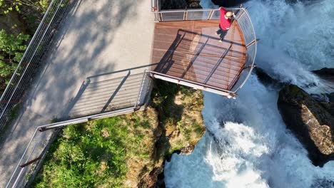 tourist overlooking geiranger majestic waterfall from a cliffside platform during summer vacation - birdseye aerial