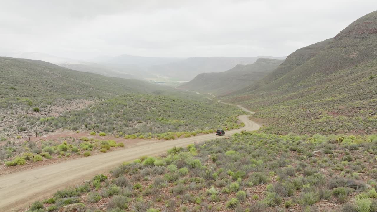 el avión no tripulado sube lentamente hacia arriba junto a una carretera donde un vehículo fuera de carretera negro conduce en el área silvestre de cederberg en sudáfrica - en el fondo se pueden ver montañas rocosas