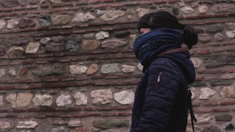 side shot of a girl in winter clothes, blue scarf and brown hair in a pony-tail as she is walking past a maroon brick wall of an old house in an old bazaar