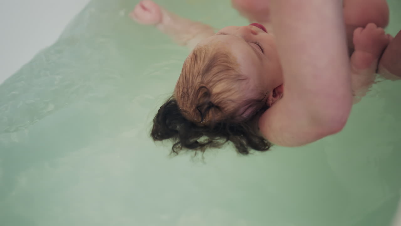 Close up of newborn baby being gently supported in bath water by caregiver during bath time near a rubber duck