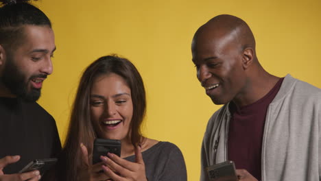 studio shot of group of friends with mobile phones celebrating winning money against yellow background